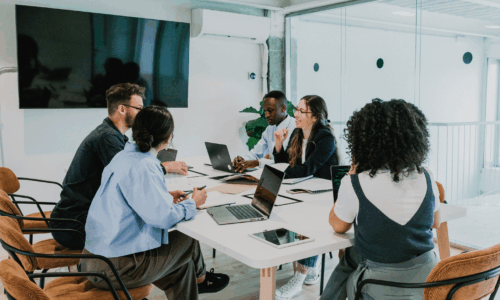 ✅ Texte alternatif (Alt text) Réunion professionnelle dans une salle moderne avec plusieurs personnes assises autour d’une table, utilisant des ordinateurs portables et discutant en groupe.