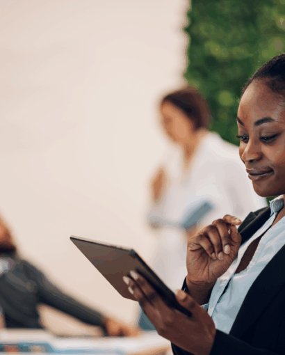 Personne en tenue professionnelle tenant une tablette numérique dans un espace de travail moderne, avec des collègues en arrière-plan discutant.