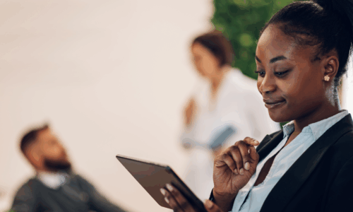 Personne en tenue professionnelle tenant une tablette numérique dans un espace de travail moderne, avec des collègues en arrière-plan discutant.
